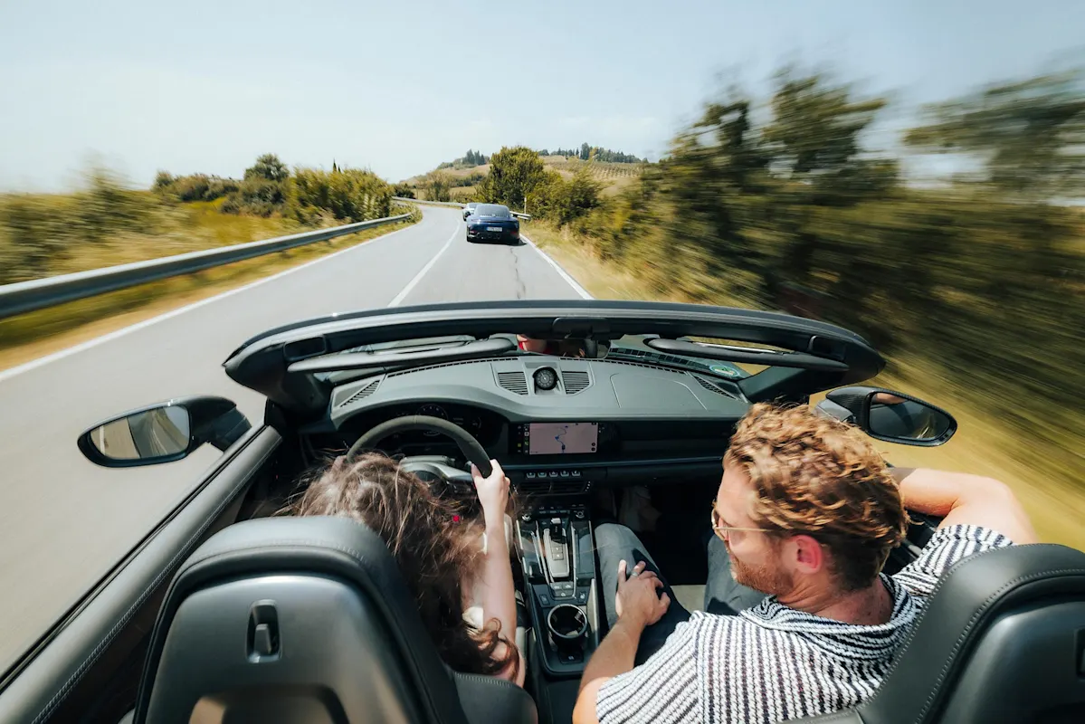 Zwei Personen in einem Cabrio auf Panoramastrecke folgen einem Porsche zwischen Bäumen.