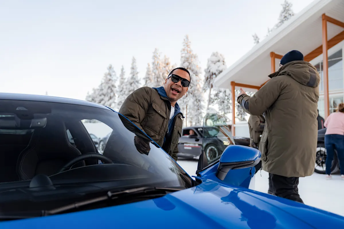 A man in sunglasses smiles beside a blue Porsche in a snowy setting with people and trees in the background.