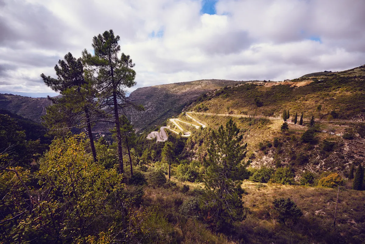 Bergstraße mit grünen Hügeln, Bäumen und teils bewölktem Himmel.