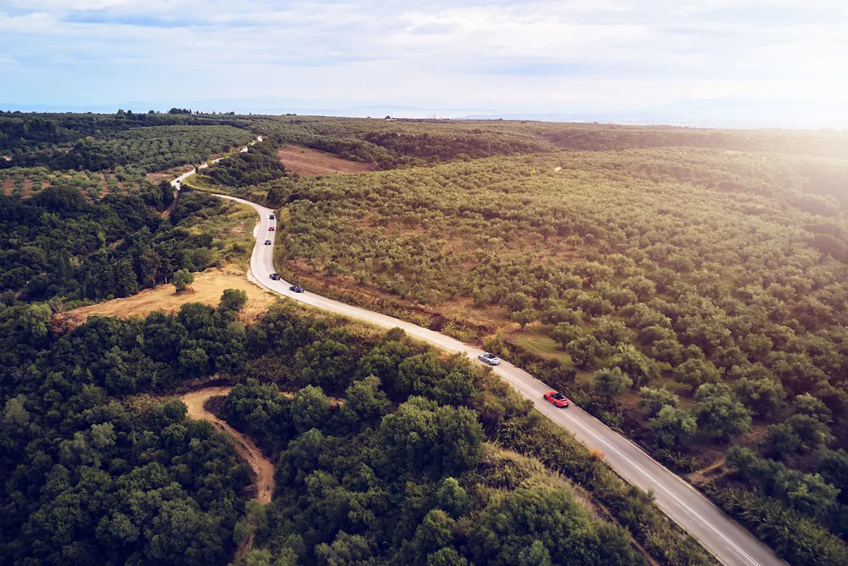 Kurvige Straße durch grüne Hügel mit Autos und Bäumen unter bewölktem Himmel.