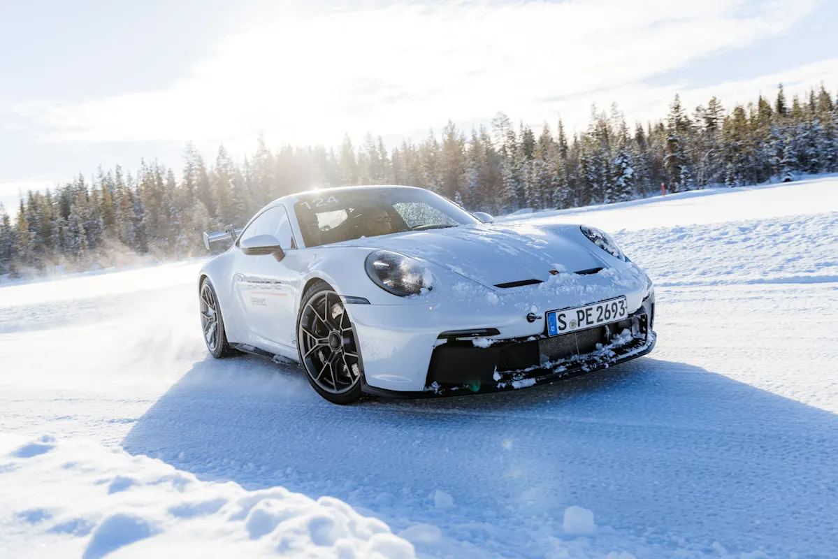 A silver Porsche speeds on snowy road, kicking up snow, with trees and sun.