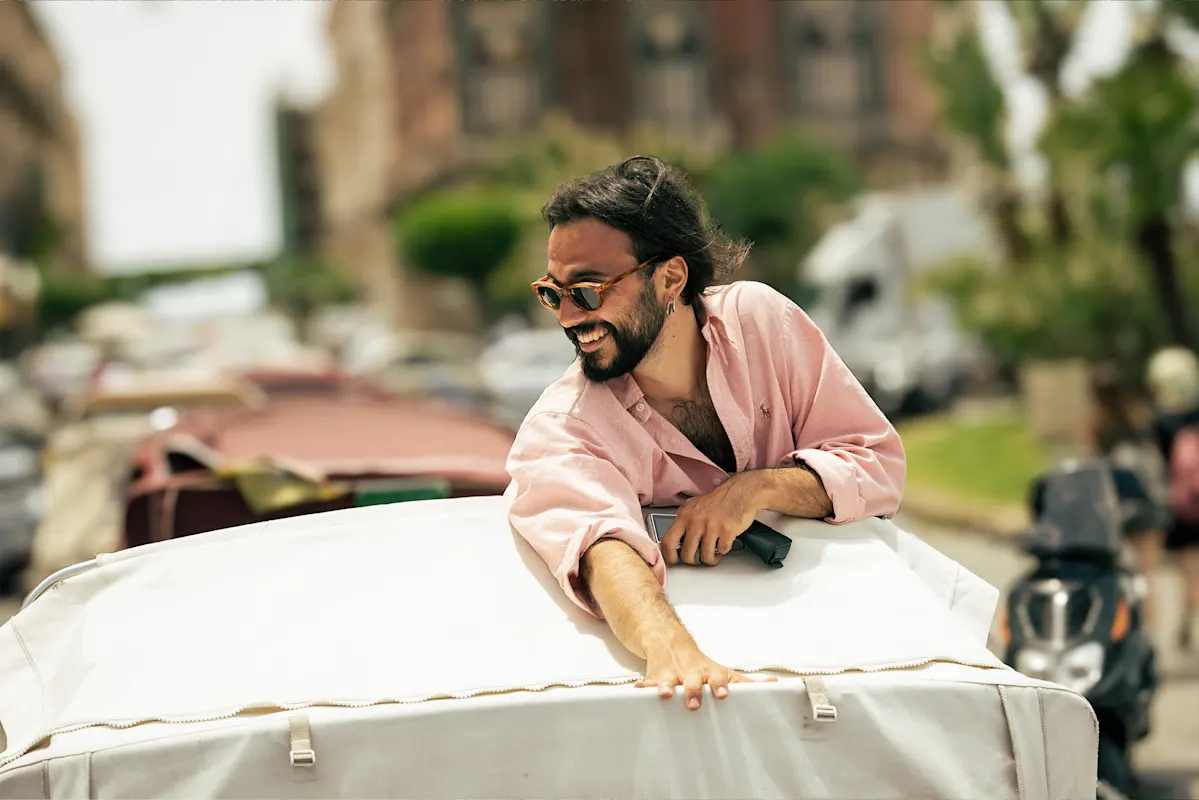 Man in sunglasses and pink shirt leaning out of a car on a sunny day.