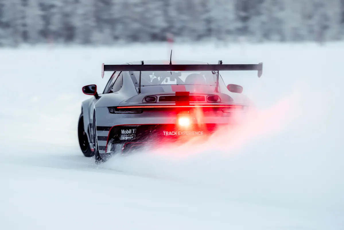 Porsche race car speeds on snowy track, red taillights glowing, kicking up snow.