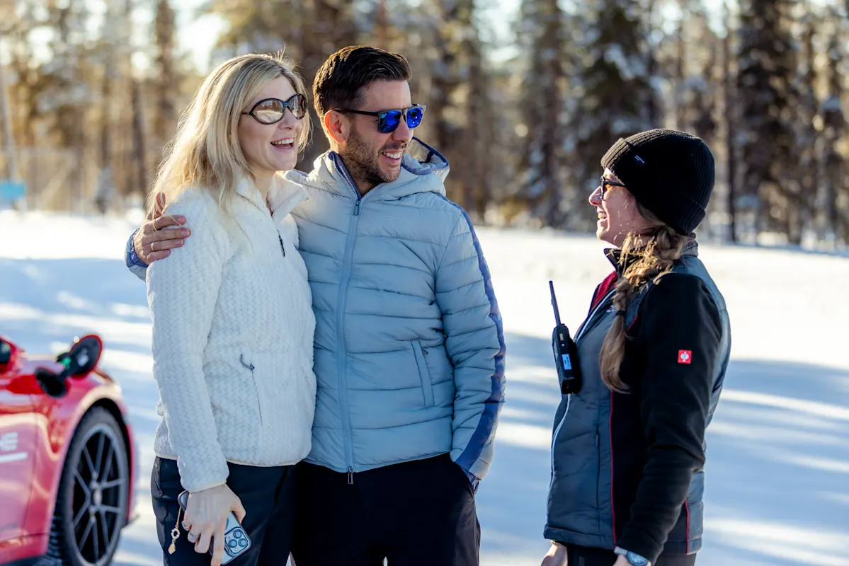 Three adults in winter clothes smile in snowy forest, with Porsche car in background.