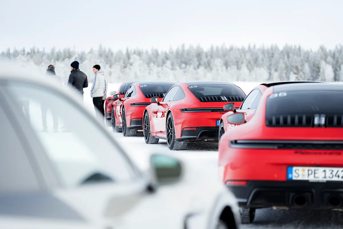 Multiple red Porsche 911s lined up on a snow track, a group of people in the background.