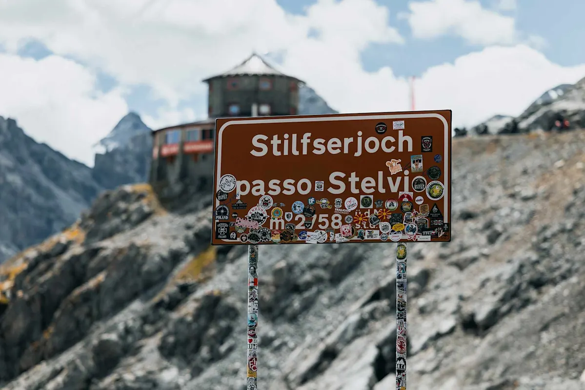 Schild Passo dello Stelvio mit Stickern, Bergen und Gebäude dahinter.