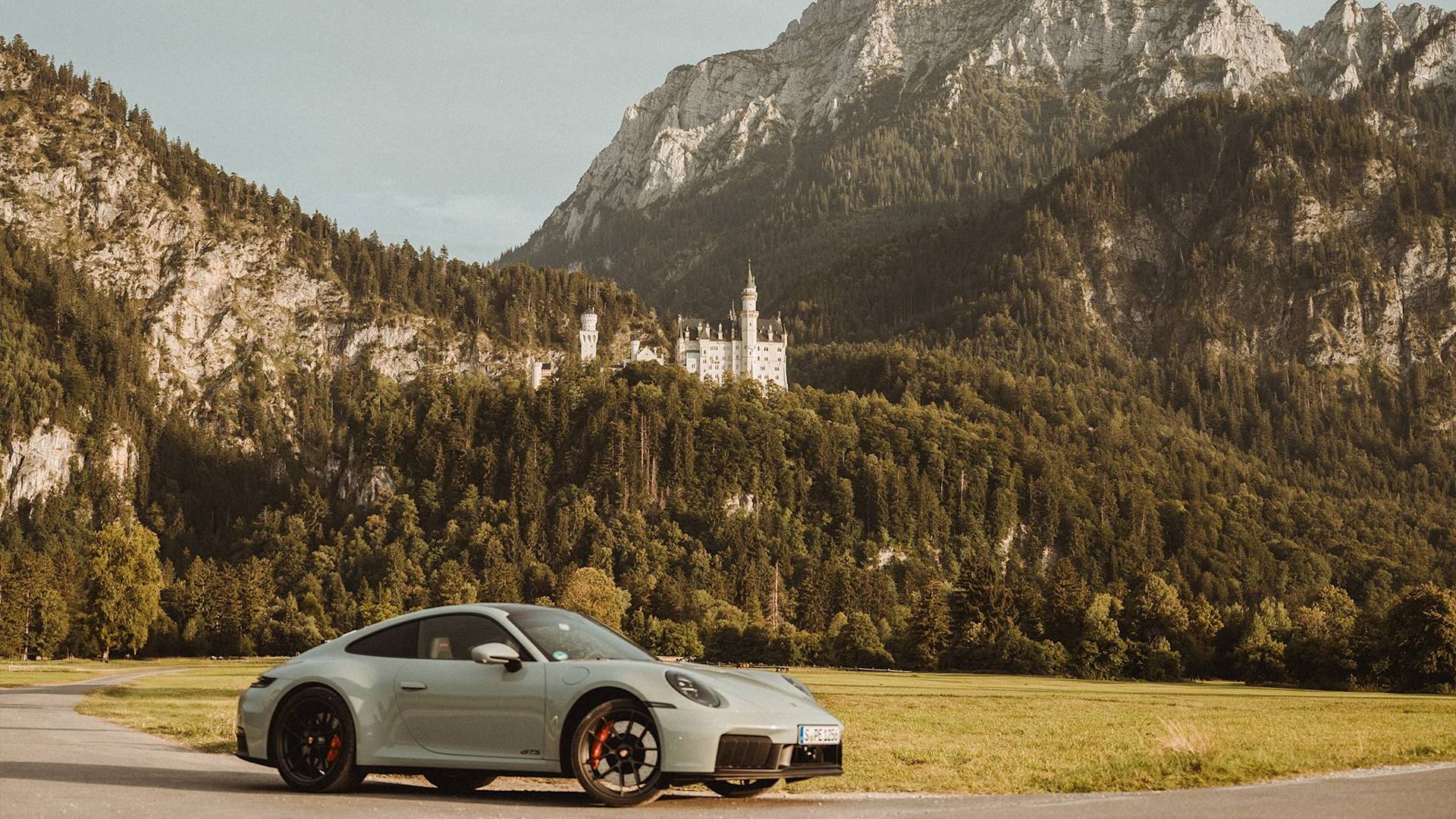 Silberner Porsche parkt auf Straße mit Burg und Bergen dahinter.