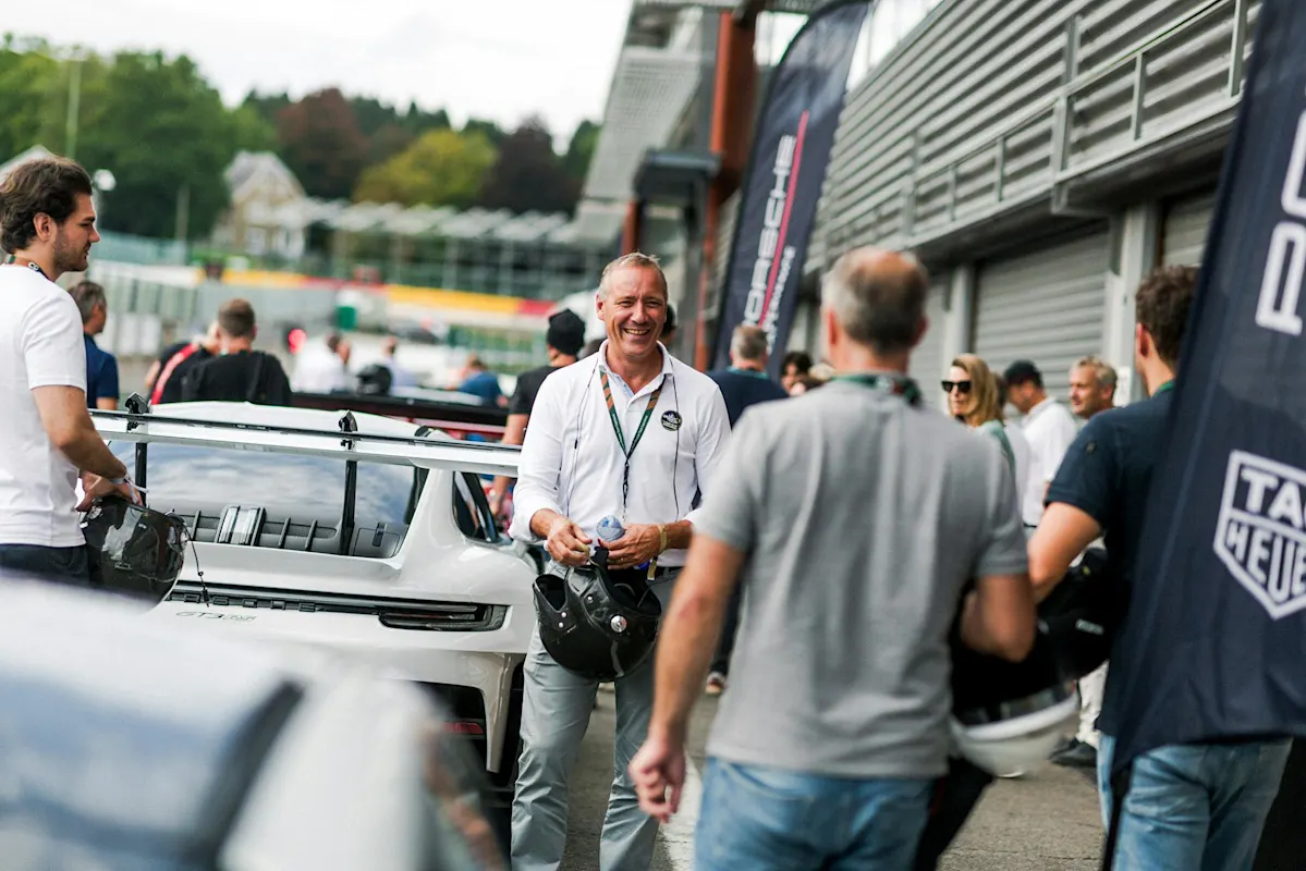 A group near a white Porsche sports car at a race event. Man in white shirt holds helmet.