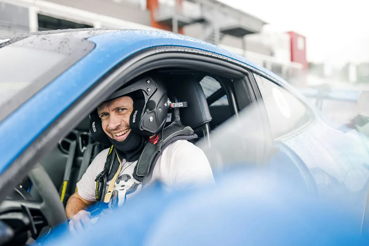 Person in helmet and racing gear, smiling in blue Porsche sports car at racetrack.