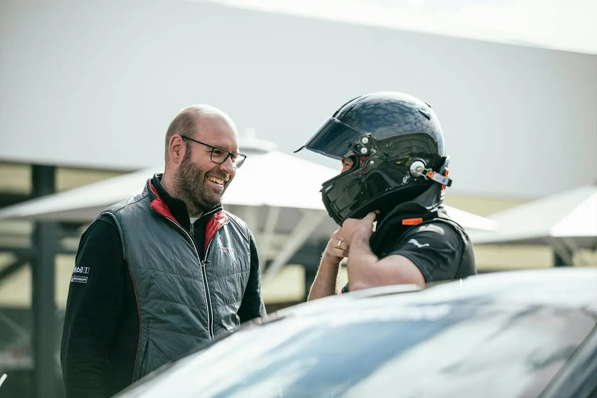 Zwei Männer, einer im Rennanzug mit Helm und der andere in einer Weste und Brille, stehen neben einem Porsche.