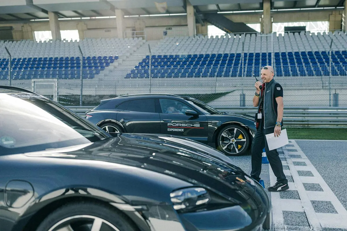 Mann am schwarzen Porsche auf der Rennstrecke, hält Papiere und Mikrofon, leere Tribünen im Hintergrund.