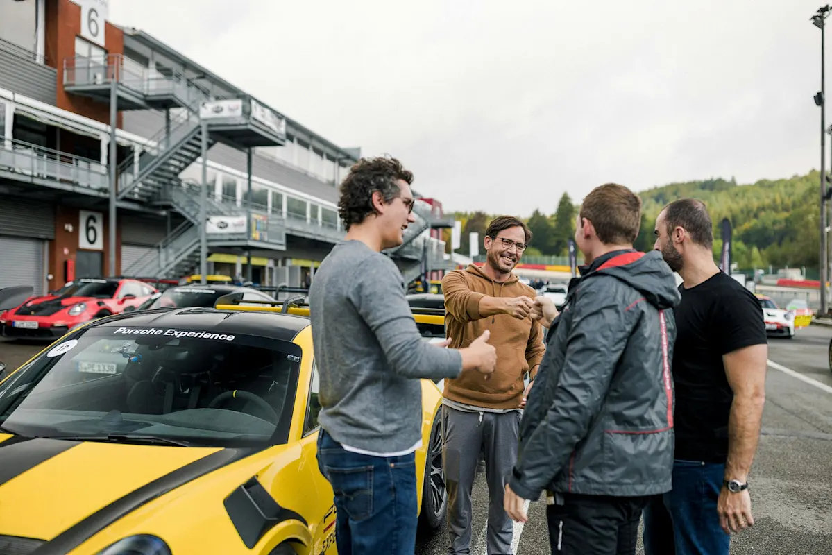 Four people laughing by yellow Porsche sports car at racetrack, with buildings and trees in the background.