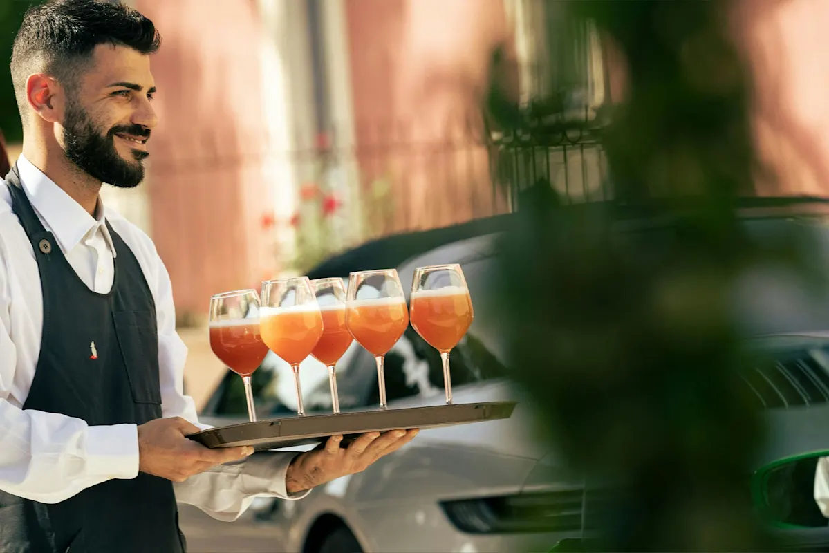Smiling server with tray of five orange drinks outdoors, blurred greenery.
