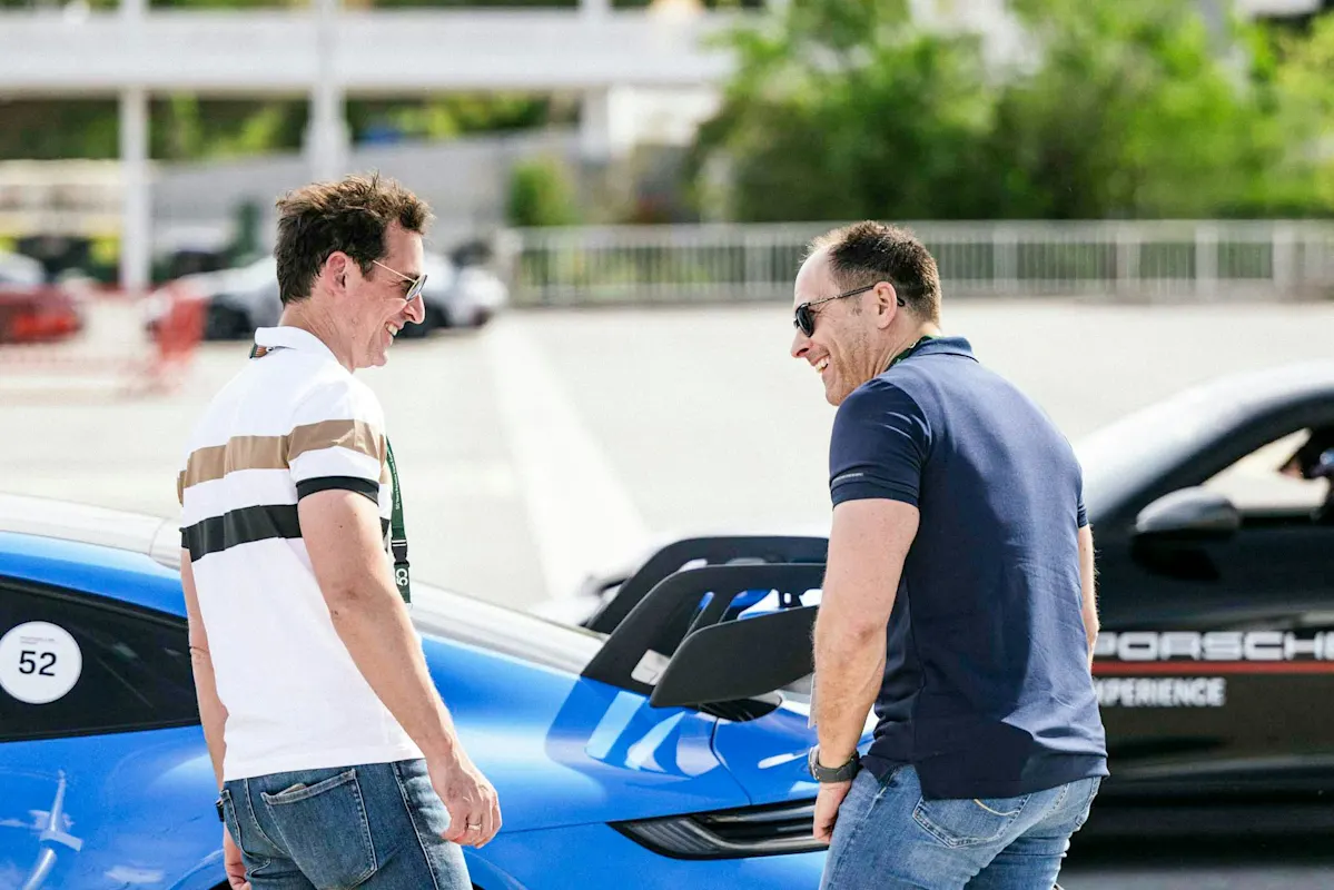 Zwei lächelnde Personen neben einem blauen Porsche Sportwagen im Freien, einer trägt eine Sonnenbrille. Beide in Freizeithemden und Jeans.