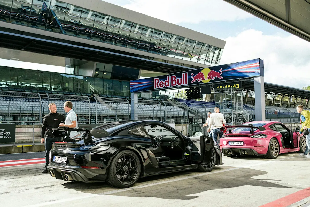 Porsche Auto in der Boxengasse an der Rennstrecke, Red Bull Schild, Menschen in der Nähe, leere Tribünen im Hintergrund.