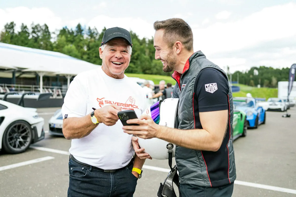 Two men chatting by sports cars on racetrack, one with helmet and phone, the other in a cap.