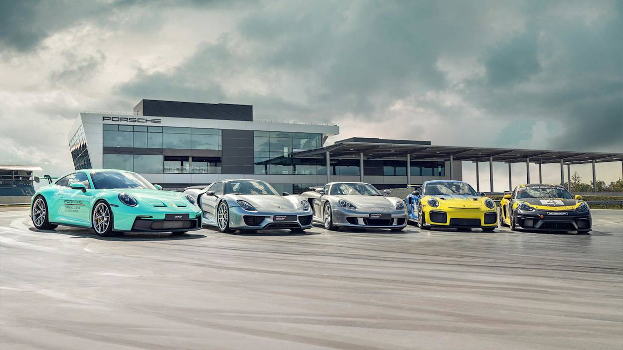 Vier Porsche Sportwagen in türkis, Silber und Gelb vor einem modernen Porsche Gebäude unter bewölktem Himmel geparkt.