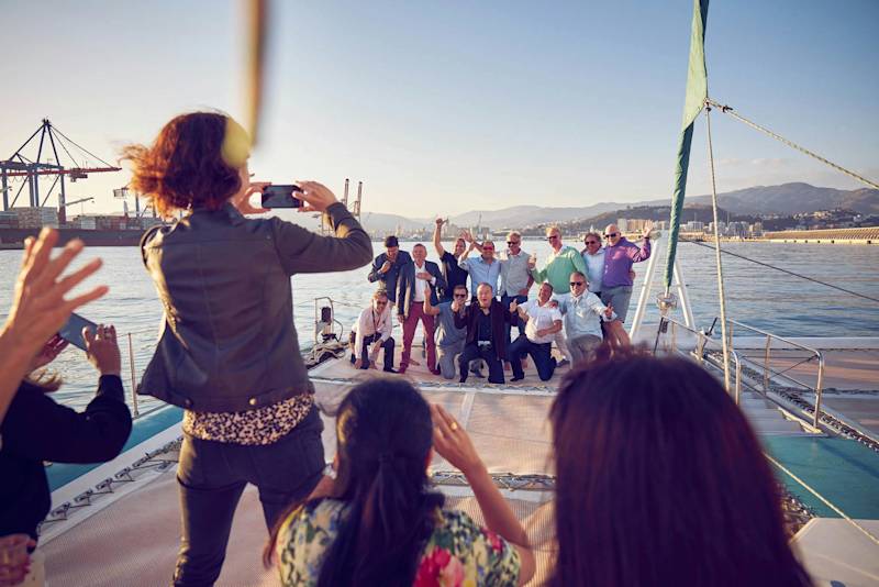 Personen posieren für ein Gruppenfoto auf einem Boot mit Stadt und Bergen im Hintergrund.