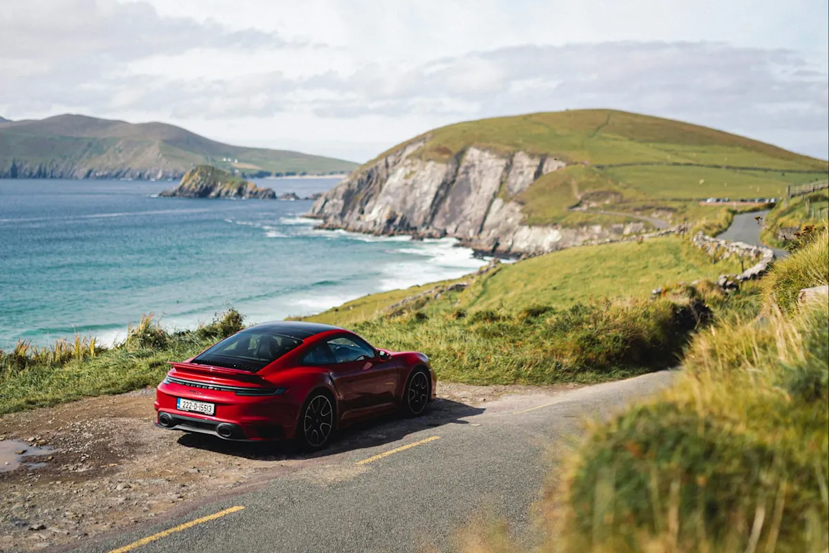 Red Porsche sports car parked on coastal road with grassy cliffs overlooking blue ocean and cloudy sky.