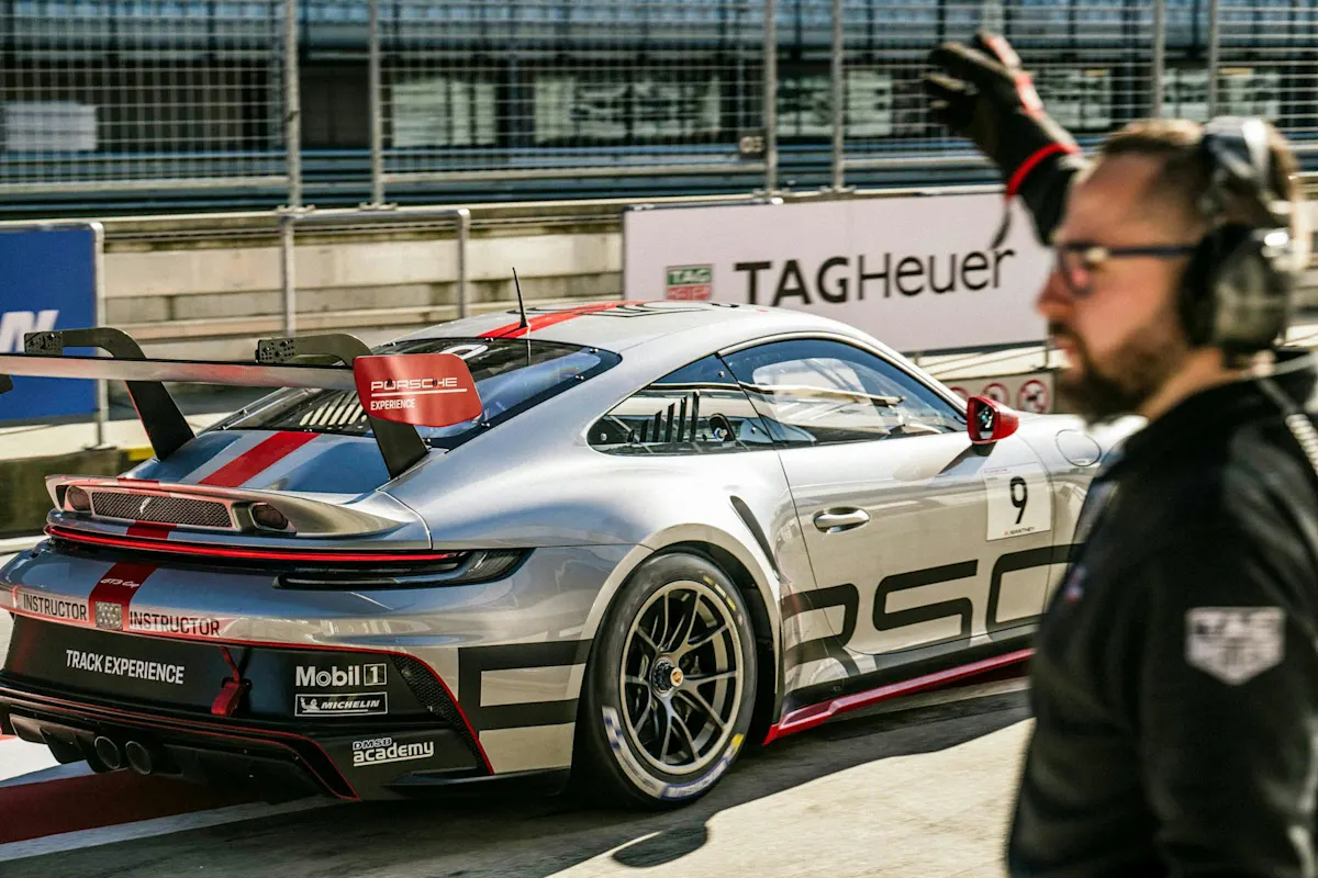 Silberner Porsche-Rennwagen mit roten Akzenten in der Boxengasse neben Person mit Headset und erhobener Hand, schwarze Jacke.