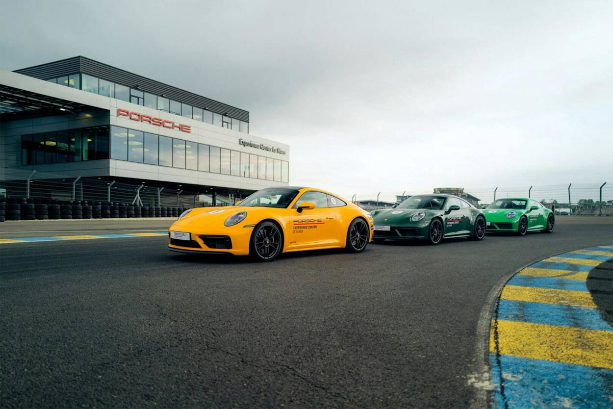 Gelber, grauer und grüner Porsche-Sportwagen auf Rennstrecke unter bewölktem Himmel mit modernem Gebäudehintergrund.