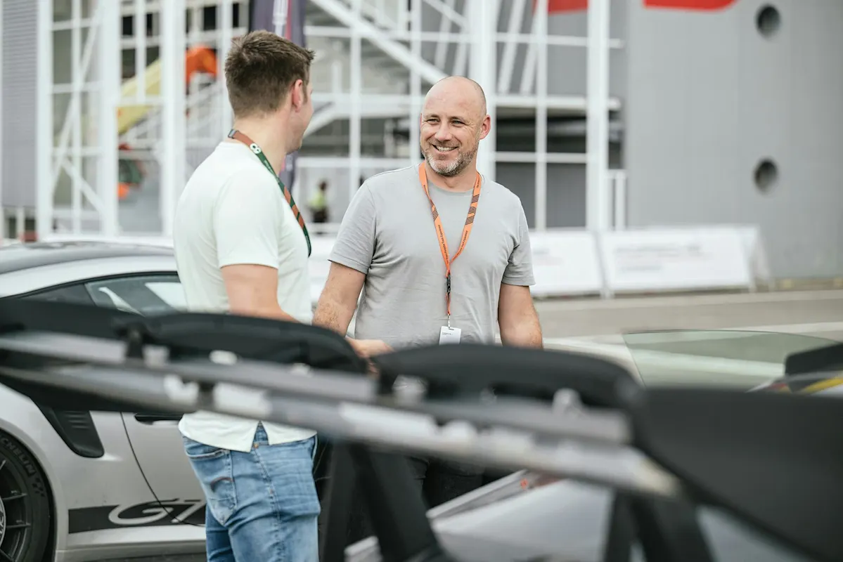 Zwei Personen lächeln neben einem Porsche Sportwagen im Freien, mit einem modernen Gebäude im Hintergrund.