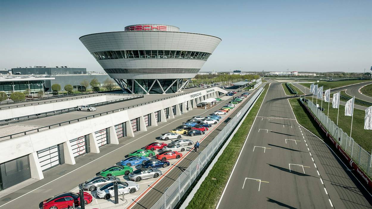 Luftaufnahme der Porsche-Rennstrecke mit farbenfrohem Sportwagen nahe der Strecke und Fahnen, unter klarem blauem Himmel.