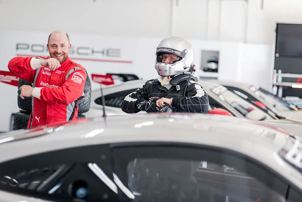 Zwei Personen in der Nähe eines Porsche-Autos - einer im Rennanzug mit Helm, der andere lächelnd und gestikulierend.