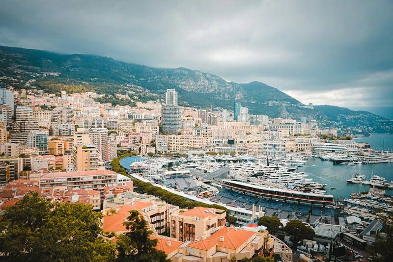 Aerial view of Monaco harbor with yachts, buildings, and mountains under a cloudy sky.