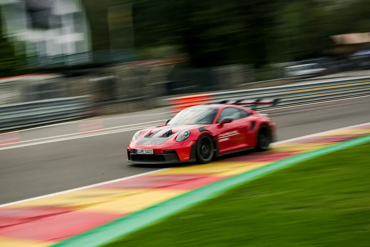 Red Porsche speeding on racetrack with blur, trees and barriers in background.