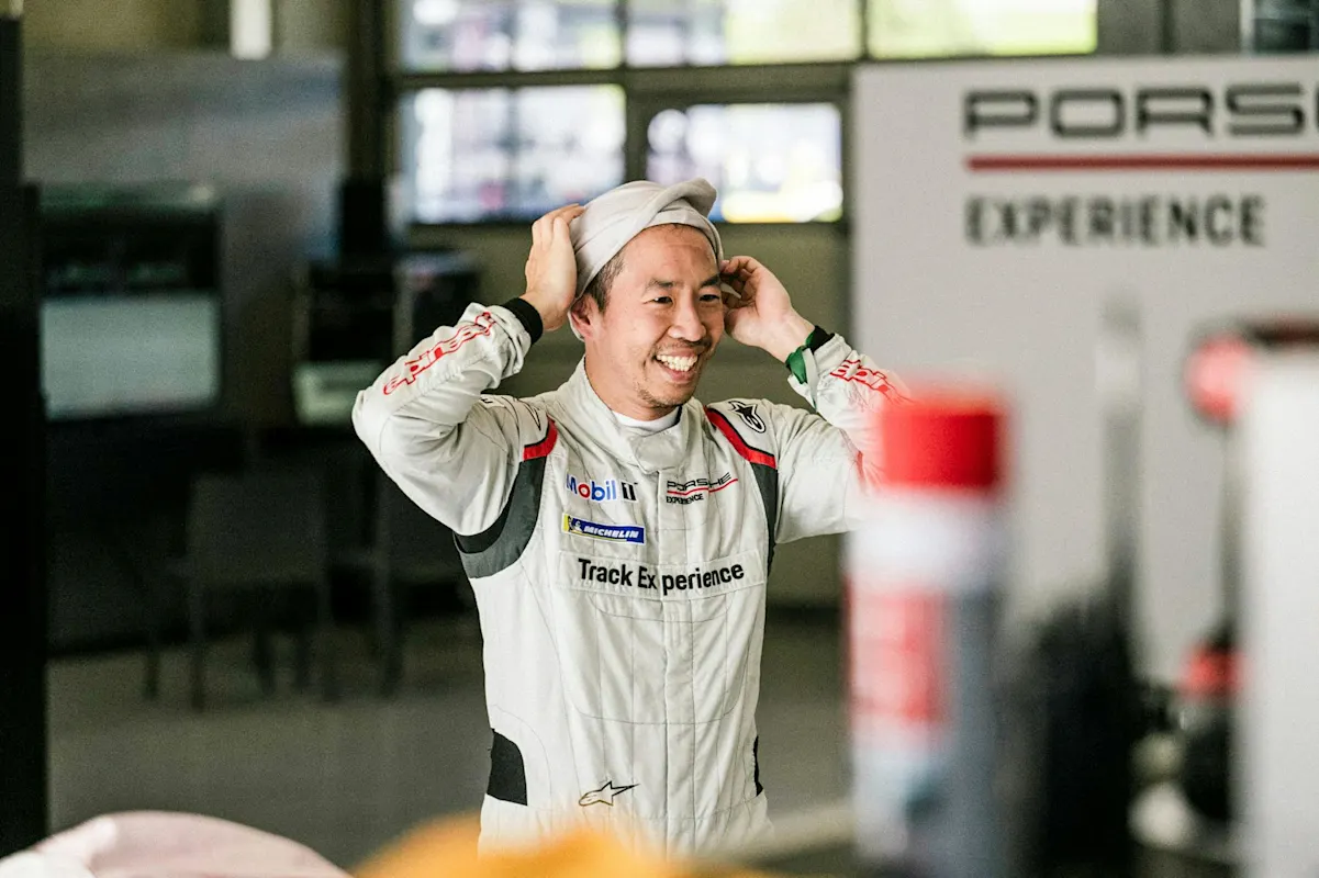 A person in a racing suit adjusts their helmet cover in a Porsche-branded motorsport garage.