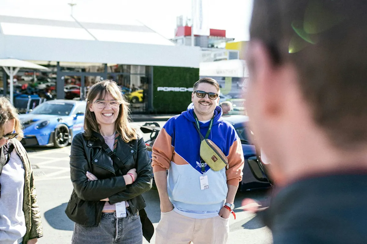 Leute lächeln neben einem Porsche, einer in Lederjacke und ein anderer in bunter Kapuzenjacke mit Umhängetasche.