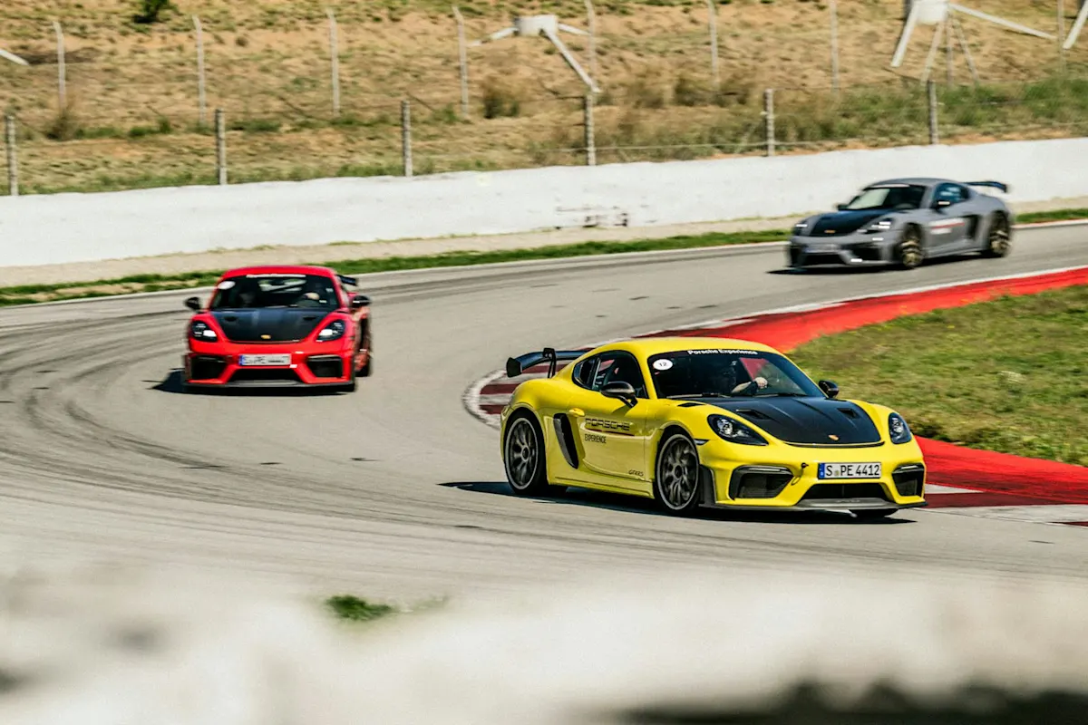 Gelber Porsche Sportwagen führt rotes und graues Auto auf der Rennstrecke entlang, umgeben von Gras und rotem Rand, unter klarem sonnigen Himmel.