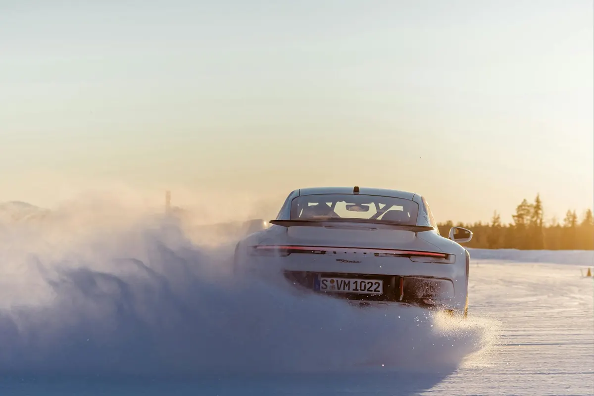 A Porsche sports car drives in snow, raising a snowy cloud, with trees in the background under a clear sky at sunset or sunrise.