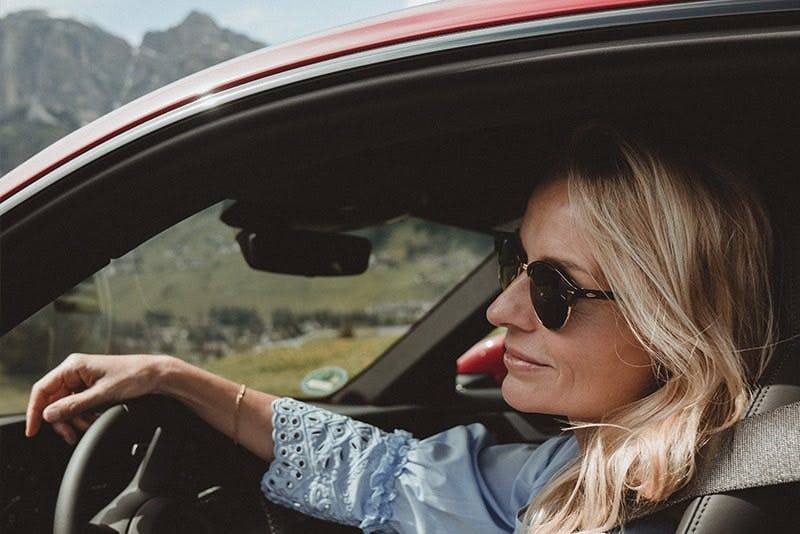 Frau fährt Porsche, lächelnd, mit sichtbaren Bergen und Grünflächen.