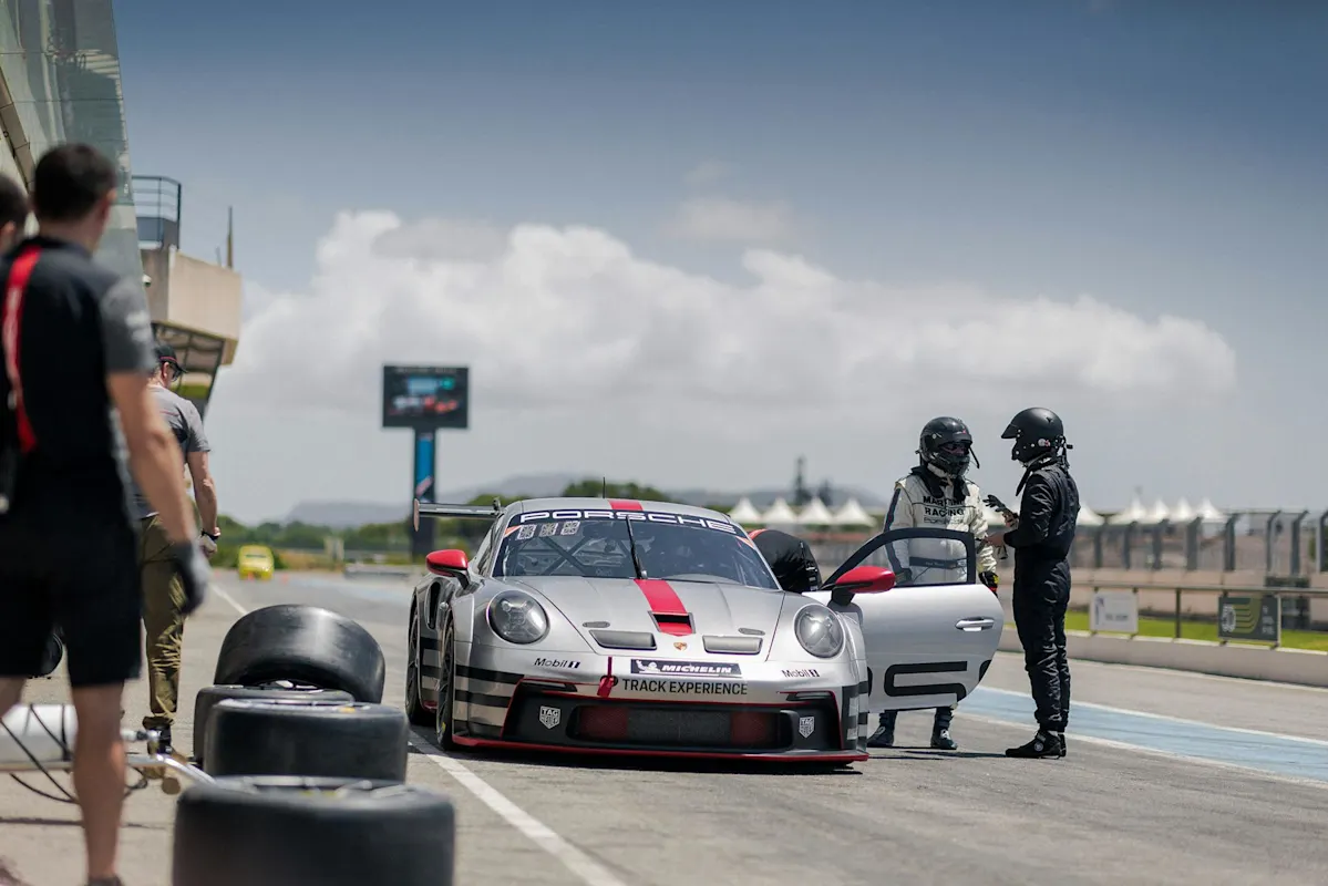 Porsche race car in pit lane with crew and spare tires nearby; cloudy sky in background.