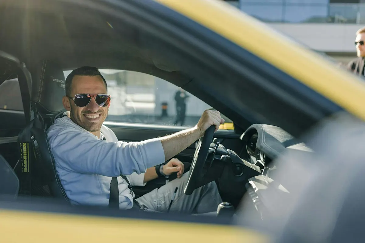Ein Mann in Sonnenbrille lächelt in gelbem Porsche und schaut in die Kamera.