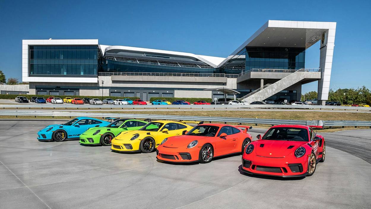 Fünf bunte Porsche-Sportwagen neben einem modernen Gebäude auf einer Rennstrecke unter klarem Himmel.