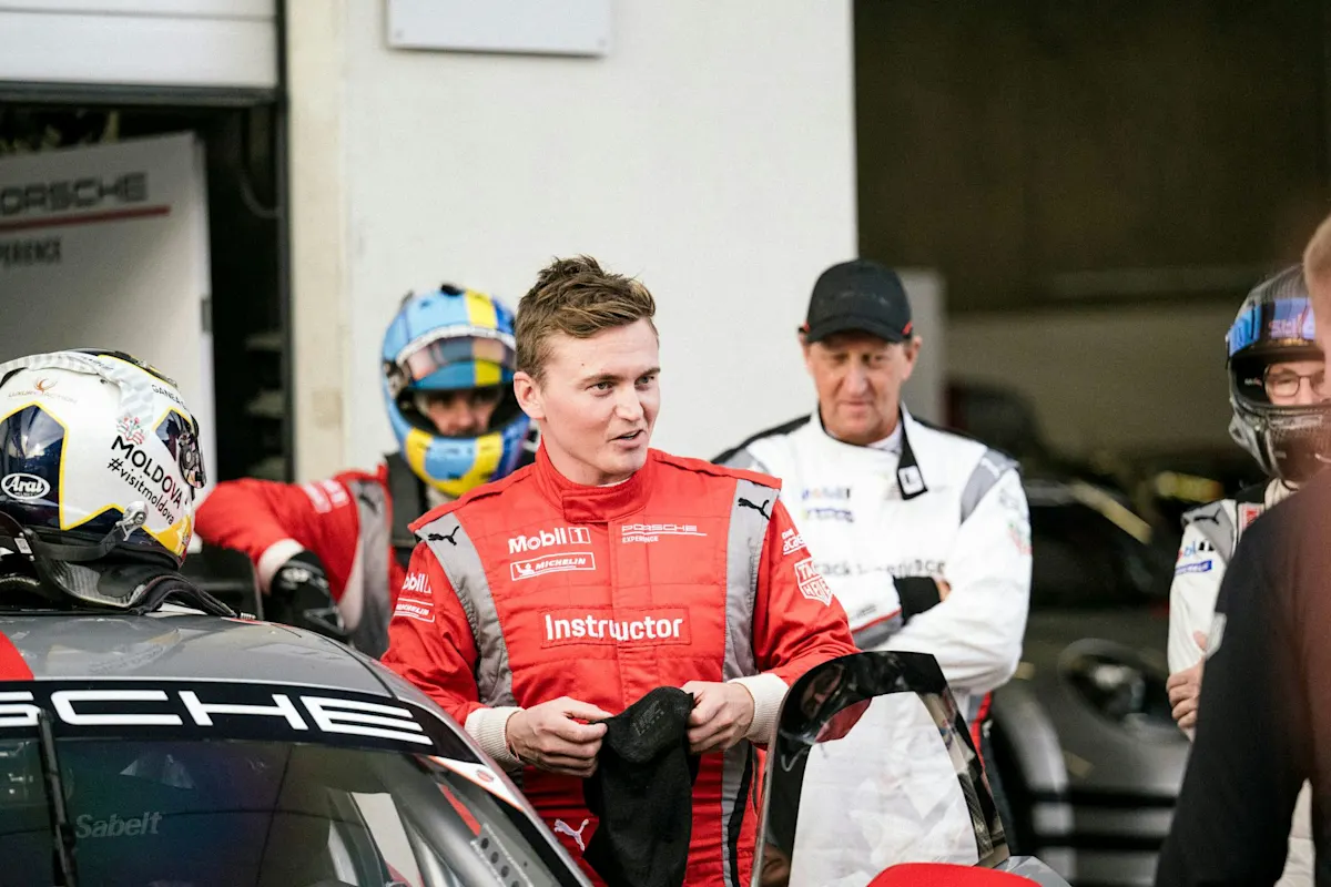 A man in a red racing suit talks to a group next to a Porsche race car. Others in helmets and suits listen.