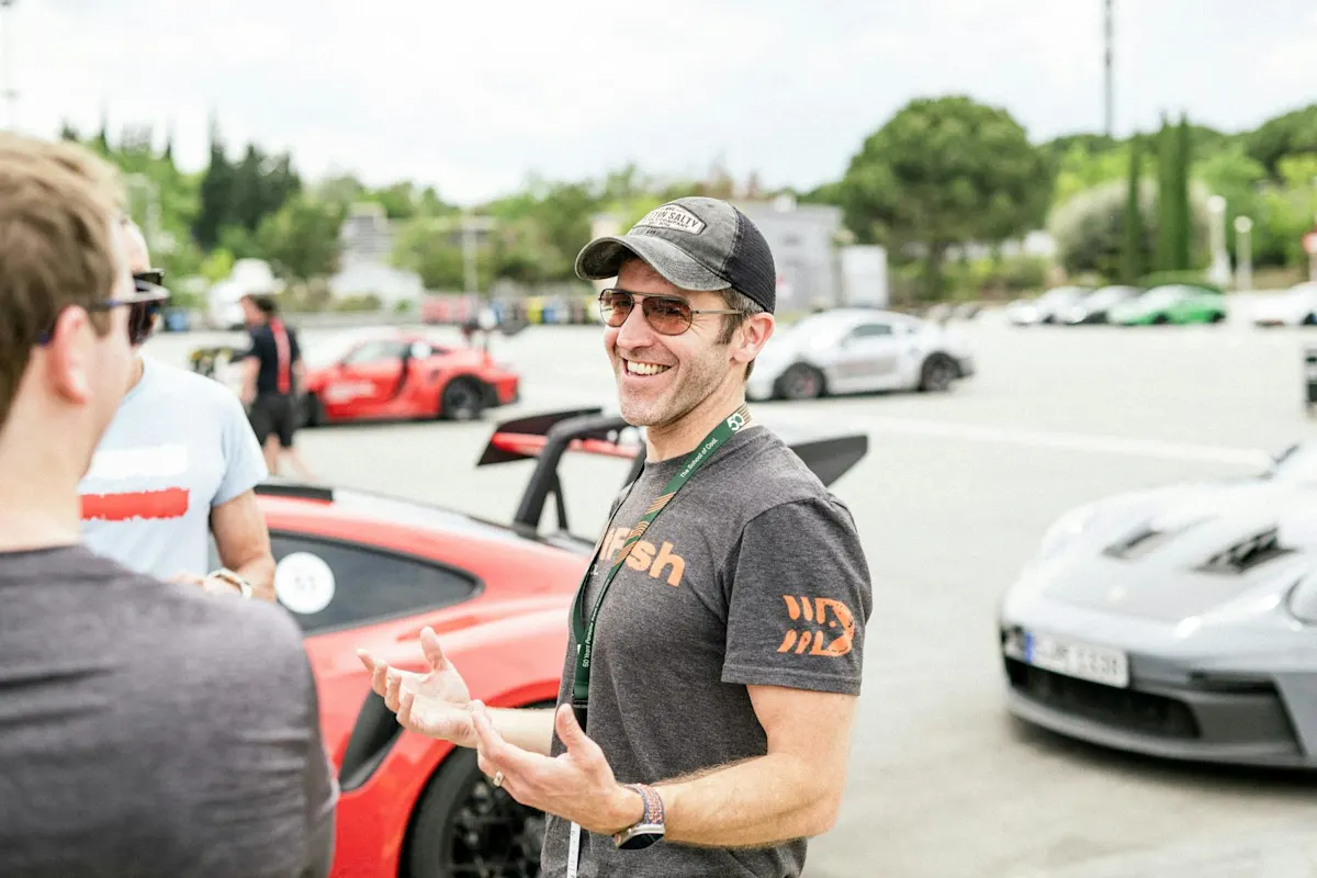 Mann in Kappe und Sonnenbrille lächelt, plaudert bei Autoveranstaltung, mit Porsche Sportwagen im Hintergrund.