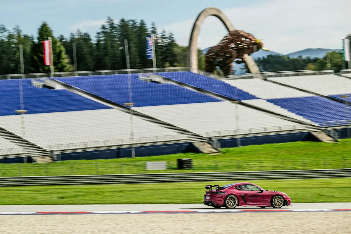 A red Porsche races on a track with a bull sculpture in the backdrop under a clear sky.