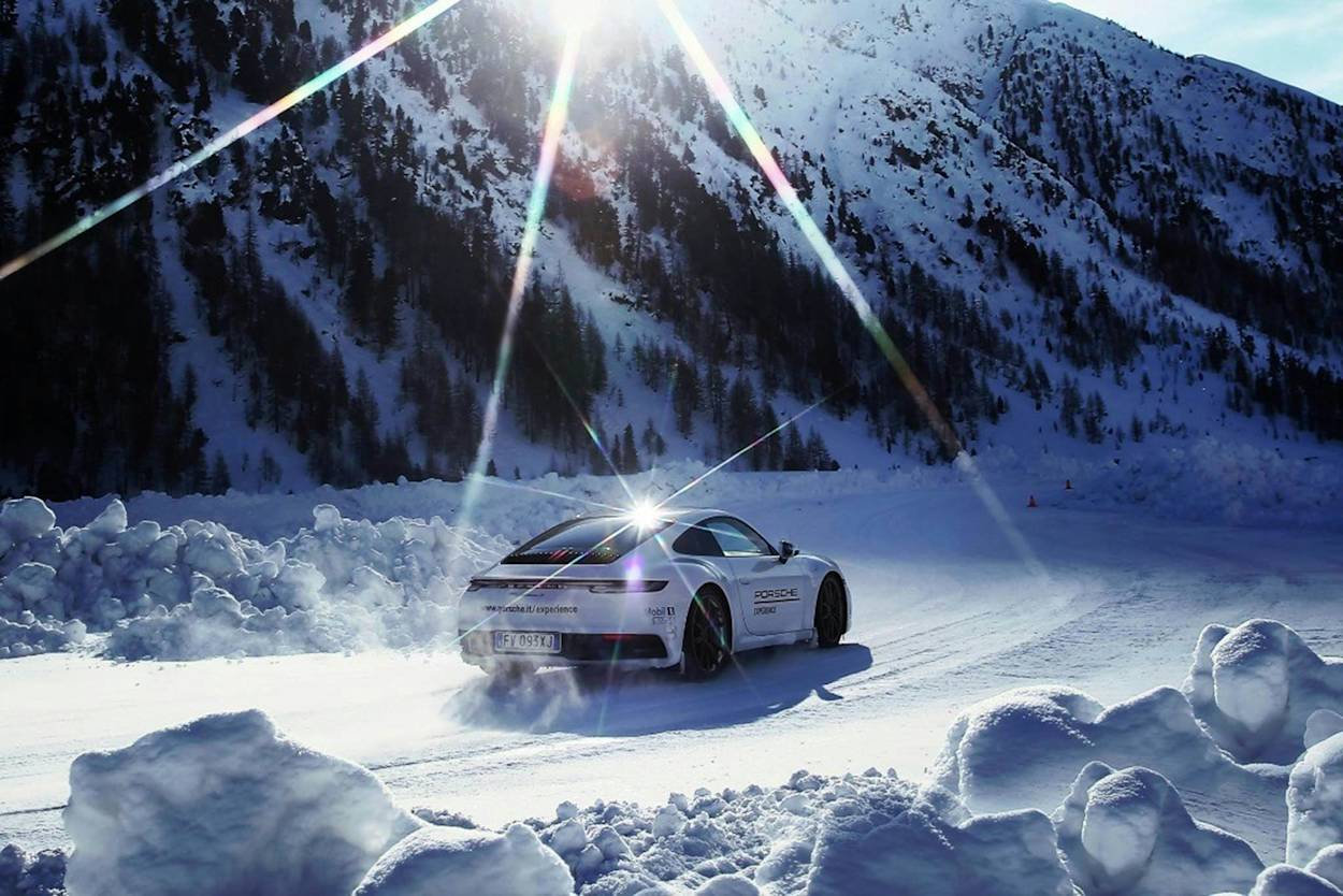 Weißer Porsche rast durch verschneite Landschaft, Sonne über Bergen, schafft kontrastreiche Szene.