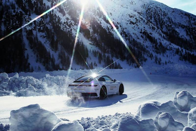 Weißer Porsche rast durch verschneite Landschaft, Sonne über Bergen, schafft kontrastreiche Szene.