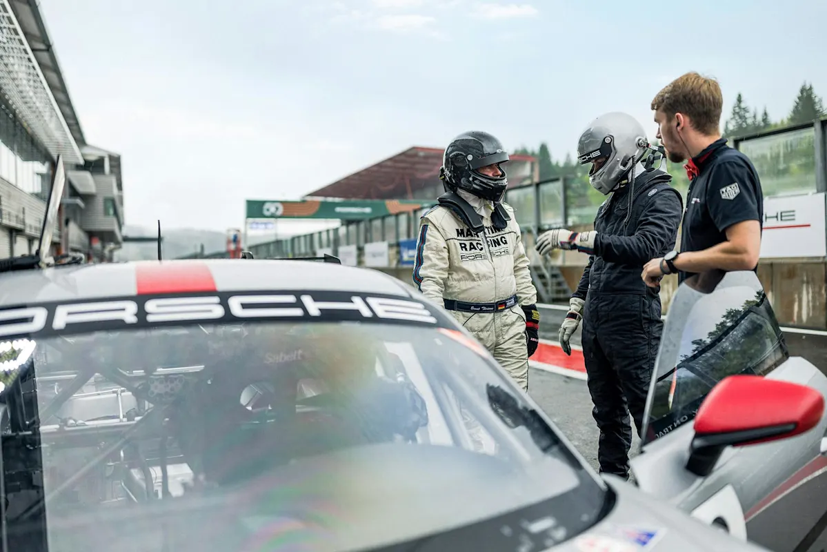 Rennfahrer im schwarzen Anzug unterhält sich mit Person im weißen Anzug neben geparktem Porsche auf der Rennstrecke.
