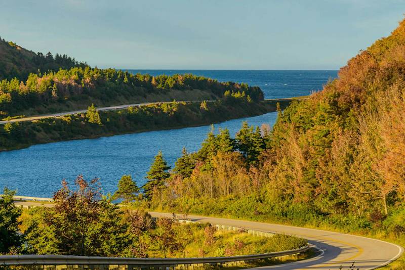 Eine kurvenreiche Straße durch üppige Bäume an einem blauen See, mit Meerwasser und klarem Himmel im Hintergrund.