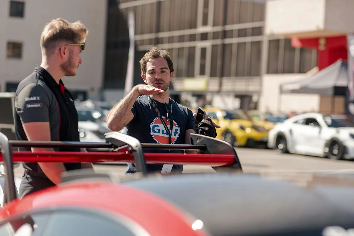 Zwei Männer neben einem roten Porsche bei einer Autoveranstaltung, mit weiteren Autos im Hintergrund.
