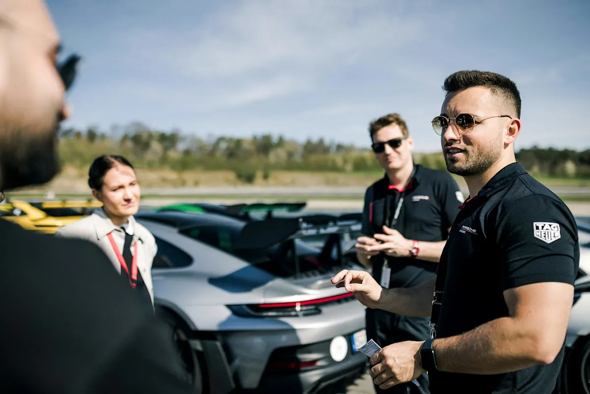 Ein Mann in Sonnenbrille spricht mit einer Gruppe neben einem Porsche Sportwagen unter klarem Himmel.