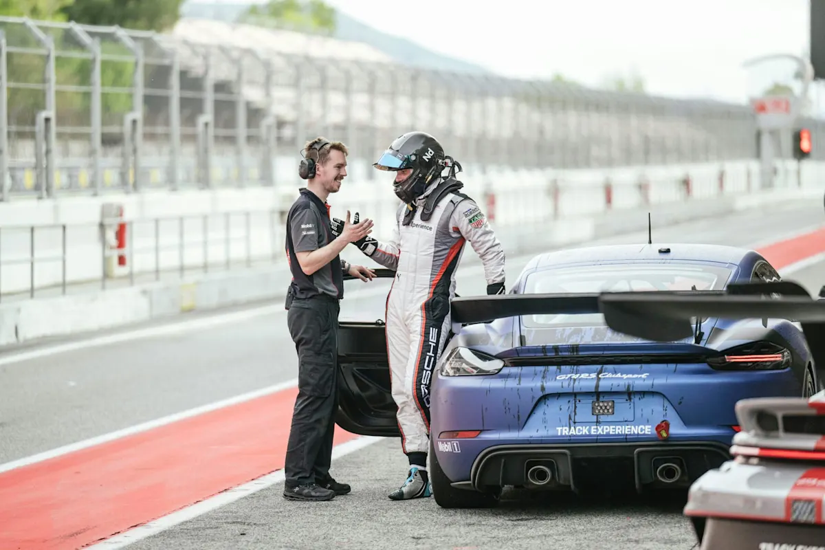 Race car driver in helmet talks to team member beside blue Porsche race car in pit lane.