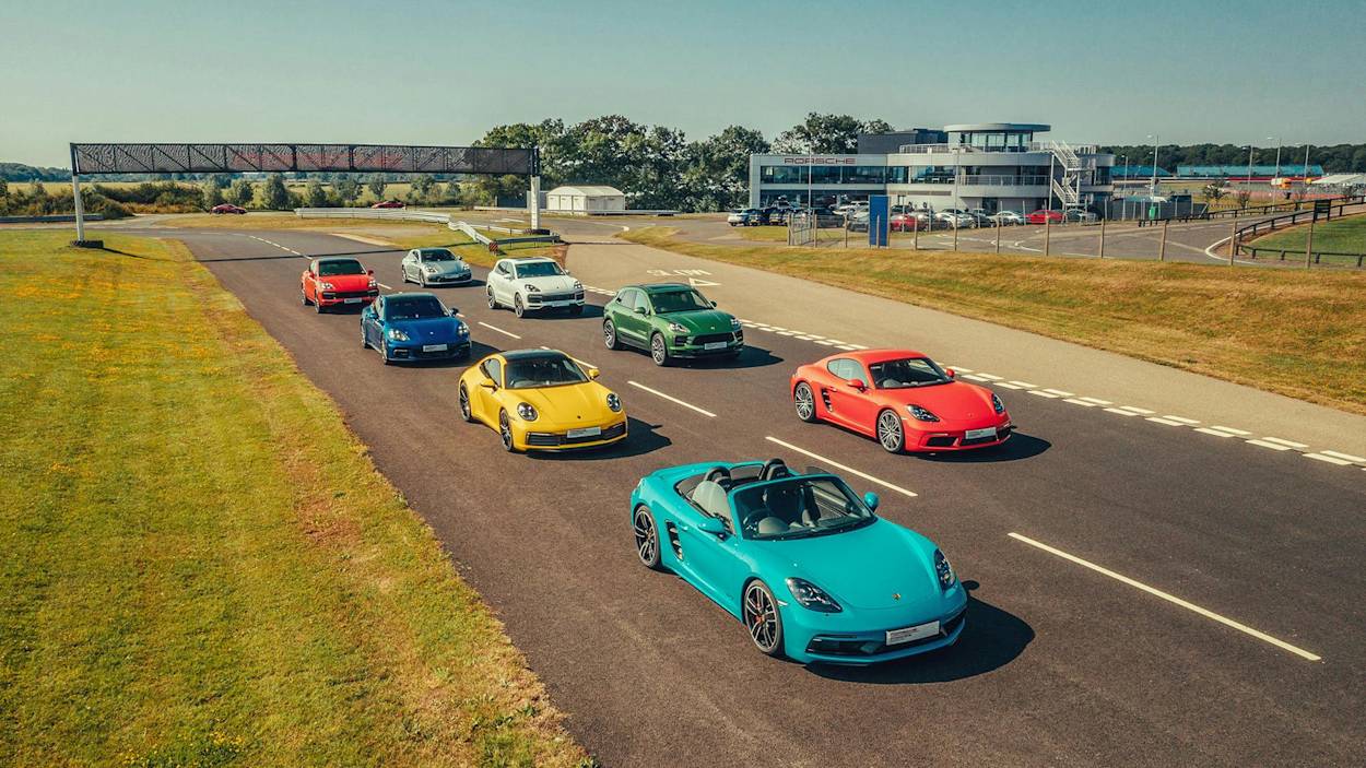 Buntes Porsche-Sportwagen-Lineup auf einer Rennstrecke, umgeben von Gras und einem modernen Gebäude unter einem klaren blauen Himmel.