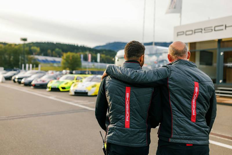 Two people in Porsche jackets embracing by parked sports cars.